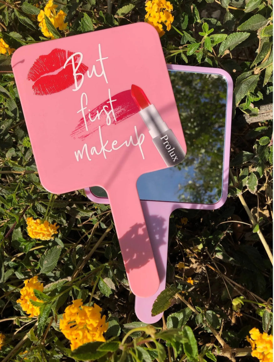 Pink hand mirror with 'But first makeup' text and lipstick graphic, surrounded by green foliage and yellow flowers.