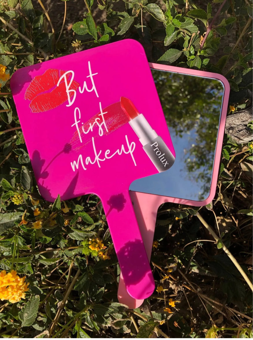 Pink handheld mirror with 'But first makeup' text and lipstick graphic on a natural background